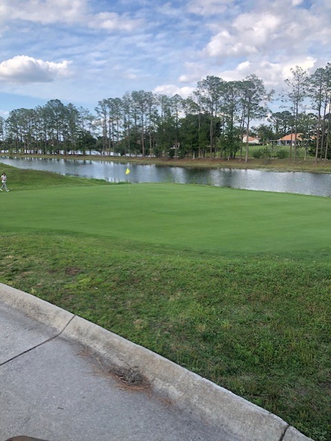 Hole 2 greens with flag, a person walking towards the flag, and trees under a bright, partly cloudy sky. A lake in the background.