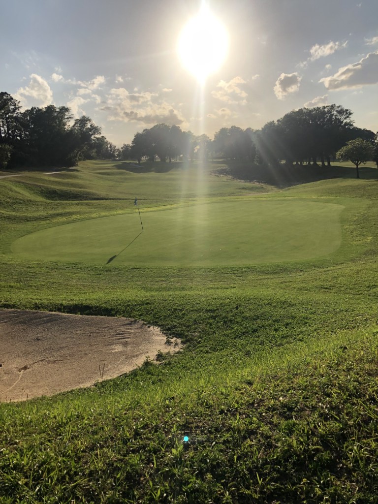 Hole 8 greens with flag under a bright, partly cloudy sky. A sand dune near the bottom left of photo.